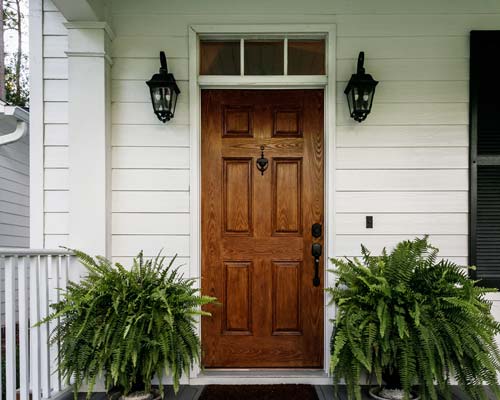 Wood six-panel entry door with transom window, black lantern sconces, and fern planters
